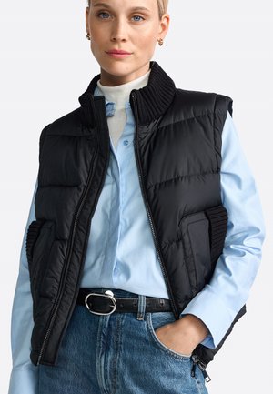 Schwarze Steppweste mit hohem Kragen, Reißverschluss und gerippten Akzenten. Kombiniert mit einem hellblauen Hemd mit Knöpfen und Jeans.