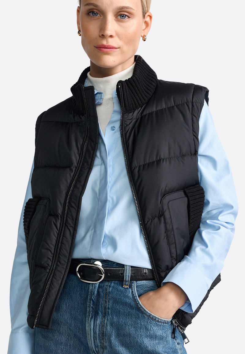 Schwarze Steppweste mit hohem Kragen, Reißverschluss und gerippten Akzenten. Kombiniert mit einem hellblauen Hemd mit Knöpfen und Jeans.