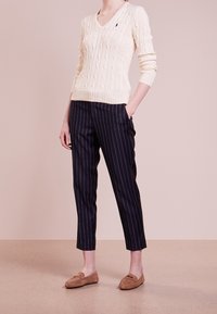 Krämfärgad kabelstickad tröja med V-hals, kombinerad med marinblå randiga byxor. Bruna mokasinskor fulländar outfiten.