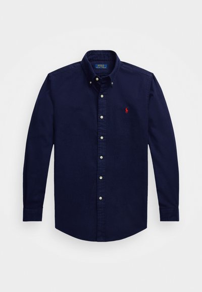 Camisa azul marinho com abotoamento em tecido suave, com gola e um pequeno logótipo bordado em vermelho no lado esquerdo do peito. Mangas compridas.