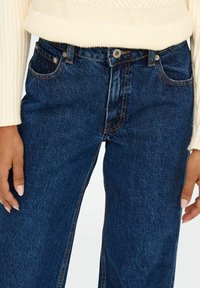 Jeans en denim bleu à taille haute et jambes larges, à la texture lisse, cinq poches et surpiqûres contrastées. Convient pour une tenue décontractée.