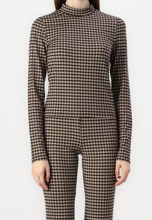Kvinde iført matchende beige og sort hundetand langærmet top og bukser mod en ensfarvet hvid baggrund.