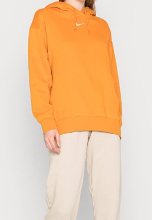 Personne portant un sweat à capuche Nike orange vif et un pantalon beige clair sur un fond uni.