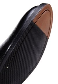 Chaussure en cuir noir avec une semelle en caoutchouc texturé ; bout arrondi et un accent en cuir marron au talon. Couture visible le long du bord.
