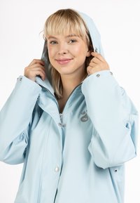 Giacca impermeabile blu chiaro con zip e chiusura a pressione, cappuccio regolabile e tasche laterali. Presenta il logo sul petto. Tessuto liscio.