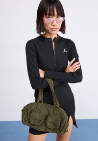 Abito nero zip-up con maniche lunghe e spacco laterale, caratterizzato da un logo bianco. Borsa in corda verde con due tasche frontali e hardware dorato.
