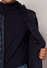 Granatowa pikowana kamizelka rozpięta na ciemnym swetrze, która eksponuje gładkie tekstury i dopasowany krój. Widoczny zamek błyskawiczny oraz umiejscowienie rąk podkreślają detal.