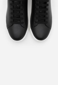 Paire de baskets en cuir noir avec lacets noirs et semelles blanches sur fond blanc, vue de dessus.