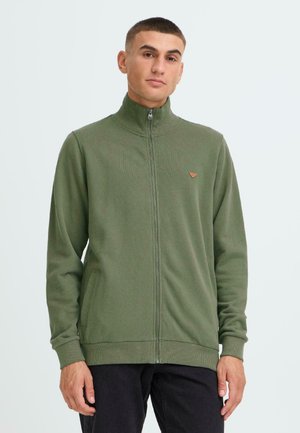 Groene ritsvest gemaakt van zachte stof met een hoge kraag, ribbelmanchetten en een klein logo op de borst. Twee zijzakken zichtbaar.