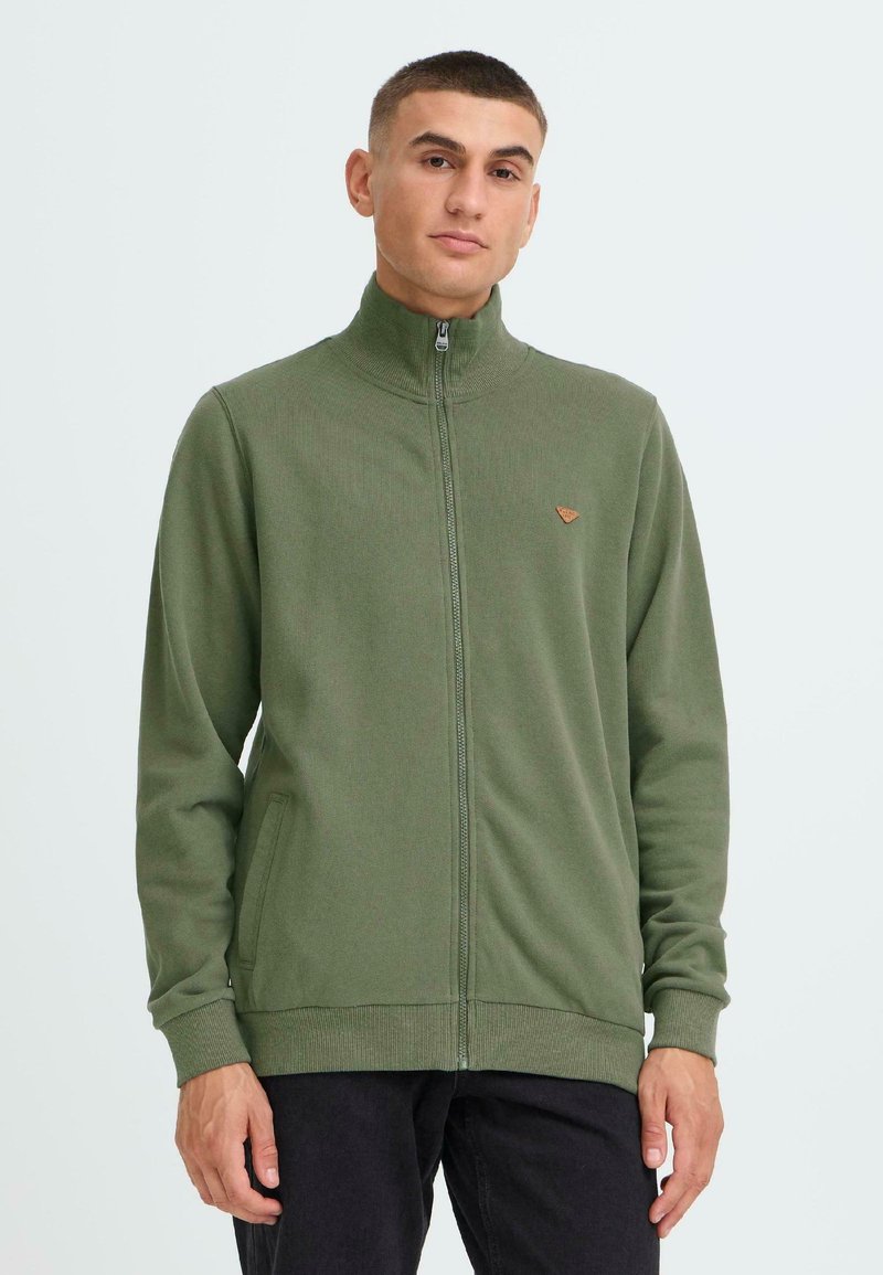 Grüne Zip-Up-Hoodie aus weichem Stoff mit hohem Kragen, gerippten Bündchen und kleinem Logo auf der Brust. Zwei seitliche Taschen sichtbar.