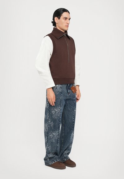Braune ärmellose Weste mit Reißverschluss über einem weißen Langarmhemd, kombiniert mit lockeren blauen Jeans aus Denim mit floraler Stickerei und einem braunen Etikett.