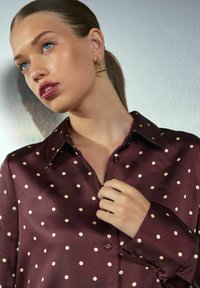 Joven mujer con ojos azules que lleva pendientes de oro y una blusa de satén color burdeos con lunares beige, sosteniendo el cuello cerca de su cuello.