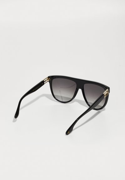 Óculos de sol pretos com lentes em gradiente, armação de plástico grossa, detalhes em metal dourado e braços das hastes curvados. Design elegante e moderno.