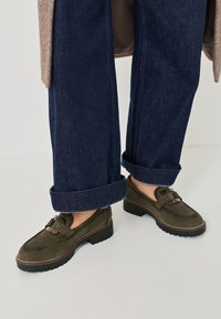 Gröna mockaloafers med en texturerad yta, prydda med en dekorativ spänne och en tjock, svart gummisula. Bärs med uppvikta denimbyxor.