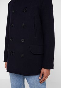 Cappotto di lana navy doppiopetto con sei grandi bottoni neri, una tasca laterale e una finitura in tessuto texturizzato.