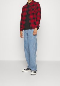 Camisa de franela a cuadros roja y negra, camiseta negra por debajo, jeans de pierna ancha de color azul claro y zapatillas negras con suelas blancas.