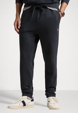 Mężczyzna ubrany w czarne spodnie dresowe z sznurkiem, szare skarpetki, białe buty sportowe z czarnymi paskami oraz zegarek na lewej ręce.