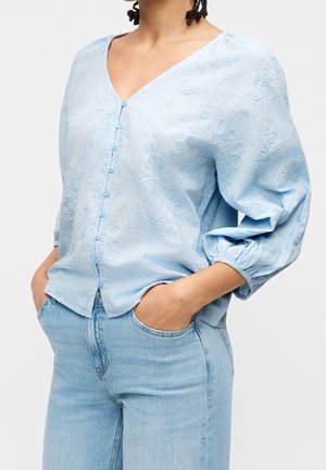 Blouse bleu clair avec broderies florales et boutons, portée avec un jean bleu clair, modèle mains dans les poches.