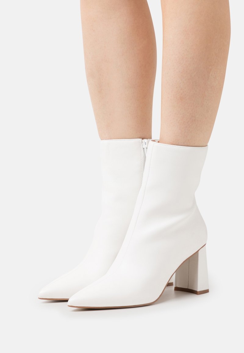 Bottes blanches en cuir montant jusqu'au milieu du mollet, avec des bouts pointus, des talons blocs et des fermetures éclair latérales, portées sur des jambes nues sur un fond uni.