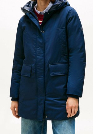 Personne portant un manteau d'hiver matelassé bleu marine avec poches à rabat, superposé à un pull rayé et une chemise à col, associé à un jean bleu.
