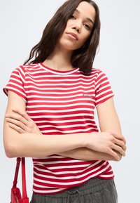 Νέα γυναίκα με μακριά σκούρα μαλλιά φοράει κόκκινη και λευκή ριγέ κοντομάνικη μπλούζα, γκρι παντελόνι και κρατάει κόκκινη τσάντα ώμου.