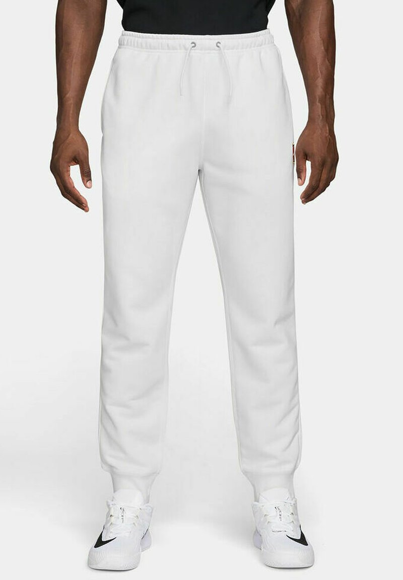 White sweatpants with a drawstring waistband, slim fit, and ribbed cuffs, paired with white athletic shoes featuring a black accent.
