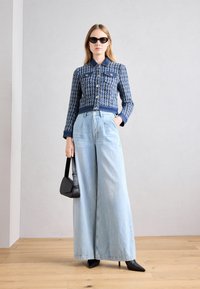 Veste courte à motifs bleus et blancs avec fermeture à boutons, associée à un jean large taille haute bleu clair et des chaussures noires à talons pointus.