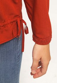 Blouse rouge à manches longueur poignet, avec un détail en dentelle sur les poignets. L'ourlet a un détail de lien froncé. Portée avec un jean en denim bleu clair.