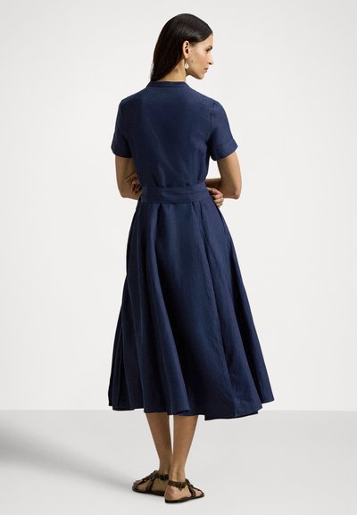Haljina od lanenog materijala tamnoplave boje s kratkim rukavima, strukirana i s raširenim suknjom. Ima pojas koji se veže i pada ispod koljena.
