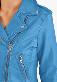 Světle modrá umělá kožená bunda se stříbrnými zipy a patentkami. Obsahuje límec s notchem a kapsy na zip. Hladká textura.