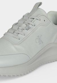Fehér Calvin Klein sneaker texturált szövettel, fűzős kialakítással és formázott talppal, melyen diszkrét márkajelzés látható az oldalon.