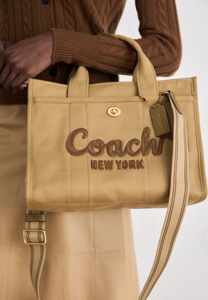 Persona con suéter marrón sostiene un bolso de lona beige Coach New York con bolsillo frontal, cierre giratorio y correa de hombro a rayas.