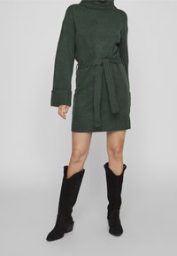 Femme portant une robe en tricot verte ceinturée avec de larges poignets et des bottes noires à bout pointu mi-mollet, debout devant un fond uni.