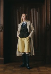 Abrigo trench beige sobre un chaleco negro acolchado, camisa blanca con cuello, pantalones cortos amarillos y botas de goma negras con calcetines de punto grises. El fondo es de madera.