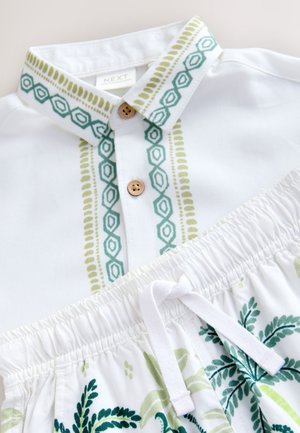 Wit overhemd met groene en turquoise geometrische borduursels langs de kraag en plooi, gecombineerd met korte broeken met bloemenpatroon en trekkoord.