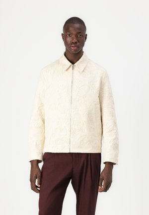 Cream quilted jacket with floral embroidery, front zipper, and a collar. Worn over dark brown pants, featuring long sleeves.