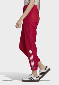 Červené sportovní kalhoty vyrobené z lehkého materiálu, se třemi bílými pruhy a logem Adidas na dolní části nohavice.