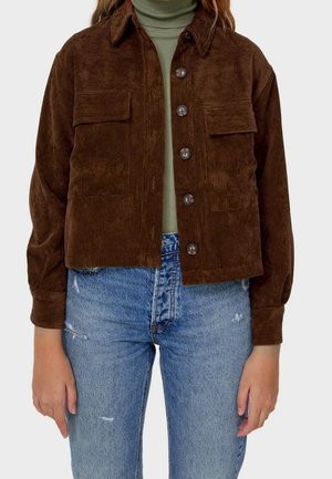 Personne portant une veste boutonnée en velours côtelé marron foncé sur un col roulé vert et un jean bleu clair déchiré.
