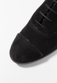 Scarpa in suede nera con una texture liscia, caratterizzata da una punta affusolata e sottili lacci neri. Accentuata da dettagli in nero lucido lungo le cuciture.
