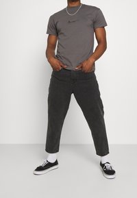 Mies, joka on pukeutunut tiukkaan harmaaseen t-paitaan, löysiin mustiin capri-housuihin, valkoisiin sukkiin ja mustiin tennareihin, seisoo kädet taskuissa.