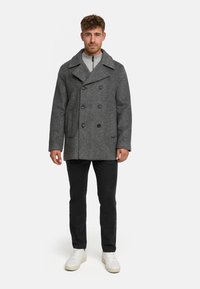 Cappotto peacoat grigio in lana con chiusura a doppio petto, ampio colletto e tasche laterali, indossato sopra una felpa leggera grigia con zip, abbinato a pantaloni neri e sneakers bianche.