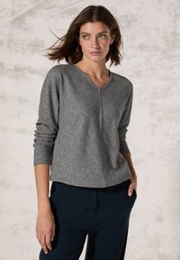 Grau gestreifter Pullover mit V-Ausschnitt und langen Ärmeln, kombiniert mit dunkelblauen Hosen, vor einem neutralen Hintergrund stehend.