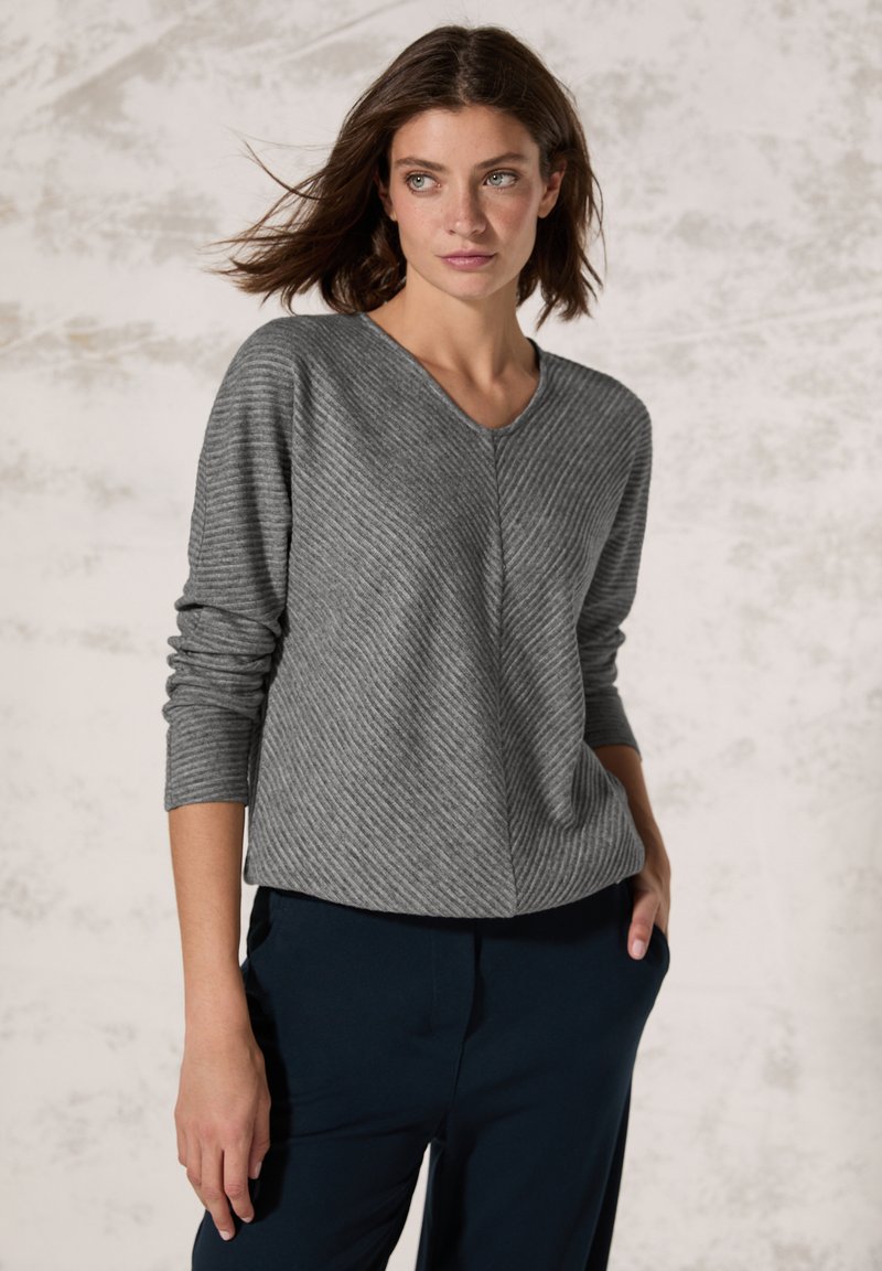 Grau gestreifter Pullover mit V-Ausschnitt und langen Ärmeln, kombiniert mit dunkelblauen Hosen, vor einem neutralen Hintergrund stehend.