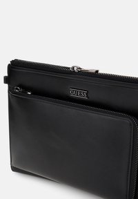 Czarna skórzana saszetka na zamek z przednią kieszenią, srebrnymi suwakami i metalową tabliczką z logo "GUESS" na gładkiej powierzchni.