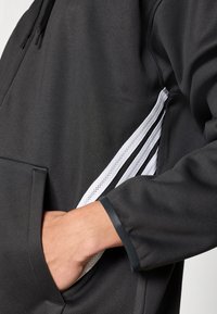 Sweat à capuche noir avec une poche avant, présentant des panneaux latéraux en maille blanche et une finition mate. Le poignet a une bordure noire contrastante.