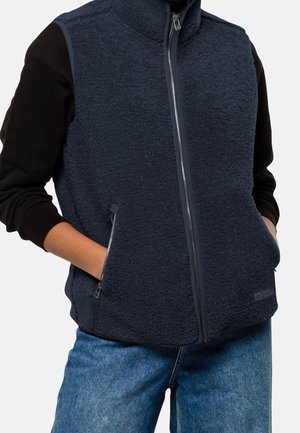 Persoon draagt een marineblauwe fleece bodywarmer met rits over een zwart shirt met lange mouwen, met handen in de borstzakken van de bodywarmer en blauwe jeans.