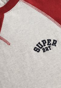 Zbliżenie na koszulkę z raglanowymi rękawami w kolorze szaro-czerwonym z czarnym haftem "SUPER DRY" na klatce piersiowej.