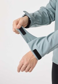 Rankos reguliuoja šviesiai mėlynos striukės rankogalius su juodu lipduku ant paprasto balto fono.