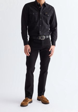Homme portant une chemise noire à boutons avec poches poitrine, pantalon noir avec ceinture et mocassins en daim marron sur fond uni.