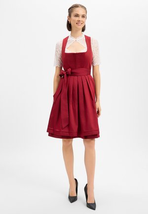 Vrouw draagt een rode geplooide jurk met een strik in de taille over een witte kanten korte mouwen blouse en zwarte hoge hakken, staand en glimlachend.